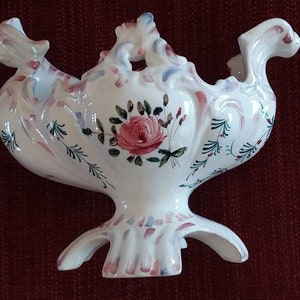 May include: A white ceramic wall pocket with a floral design. The pocket features a central rose motif, with pink, blue, and green accents. The piece has a decorative, ornate shape with a three-footed base.