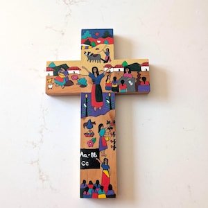 May include: A wooden cross featuring hand-painted scenes of daily life, including figures in colorful clothing, rural landscapes, and a black chalkboard section with the white text Aa-Bb Cc. The artwork depicts various activities and people.