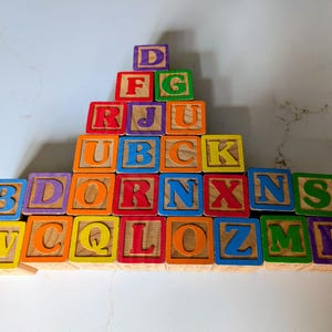 May include: A pyramid of colorful wooden alphabet blocks. Each block features a different letter in bold, uppercase font. The blocks are painted in various bright colors, including red, blue, green, yellow, and orange. The blocks are arranged on a white surface.