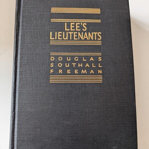 May include: A hardcover book titled "Lee's Lieutenants" by Douglas Southall Freeman. The book has a dark cover with gold lettering and horizontal lines. The cover has a textured appearance.