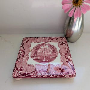 May include: Square ceramic trivet with a burgundy and white design. The trivet features a central image of a building and surrounding leaf pattern. The trivet has small white feet. A pink flower in a silver vase is in the background.