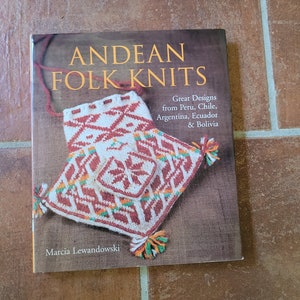May include: A book titled "ANDEAN FOLK KNITS" with a cover featuring a detailed, colorful textile design. The cover art showcases a geometric pattern in red, white, and orange, with tassels. The book's subtitle reads "Great Designs from Peru, Chile, Argentina, Ecuador & Bolivia."
