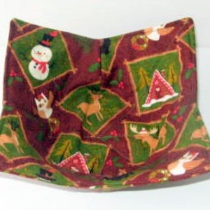 Puede incluir: Un posavasos de tela marrón y verde con temática navideña. El posavasos presenta una variedad de diseños de invierno, incluyendo un muñeco de nieve, un ciervo, un alce, una cabaña y un árbol de Navidad.