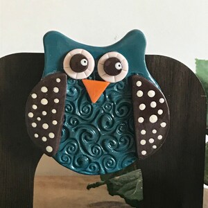 May include: A teal blue owl figurine with white polka dots on its wings. The owl has brown eyes and an orange beak. The owl is mounted on a dark brown wooden surface.