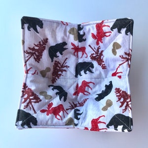 May include: White cotton pot holder with a woodland pattern featuring black bears, moose, and trees. The pot holder is quilted and has a square shape.