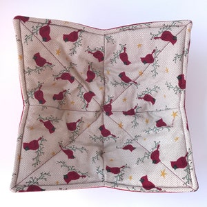 Peut inclure: Un dessous de plat carré matelassé avec un fond crème et un motif répété de cardinaux rouges perchés sur des branches vertes avec des étoiles dorées. Le dessous de plat a une bordure rouge.