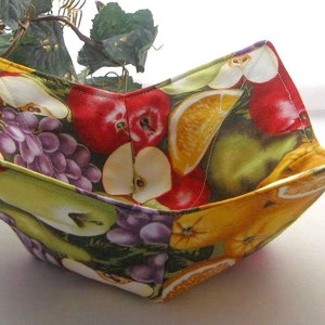 Puede incluir: Un cuenco de tela con un diseño de estampado de frutas. El cuenco presenta manzanas, uvas, peras y naranjas en tonos rojos, verdes, amarillos y morados.