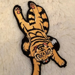 May include: A black and orange embroidered patch of a tiger with its paws in the air, looking down with a fierce expression.