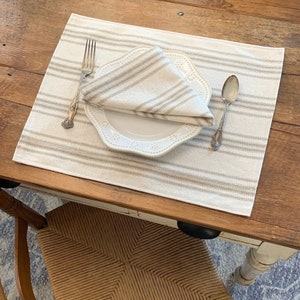 May include: A white placemat with gray stripes, a white plate with a scalloped edge, a silver fork, and a silver spoon. The placemat is on a wooden table.