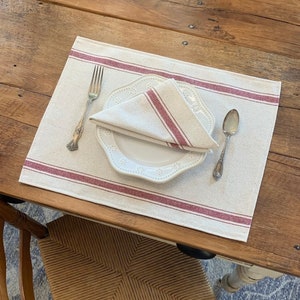 May include: A white linen placemat with red stripes, a white plate, a silver fork, and a silver spoon. The placemat is set on a wooden table.