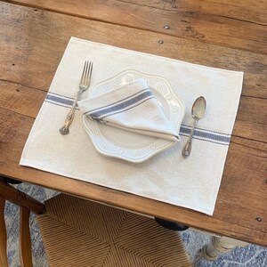 May include: A white linen placemat with a blue stripe border, a white plate with a scalloped edge, a silver fork, and a silver spoon. The placemat is on a wooden table.