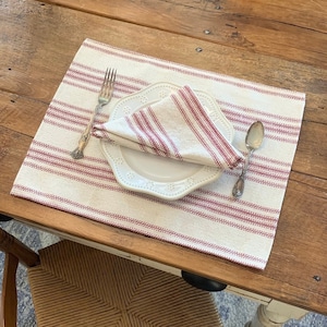 May include: A place setting on a wooden table. A white plate with a scalloped edge holds a folded red and white striped napkin. A fork and spoon flank the plate. The place mat is cream-colored with red stripes.