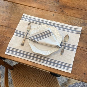 May include: A white placemat with blue and gray stripes, a white plate with a scalloped edge, a silver fork, and a silver spoon. The placemat is on a wooden table.