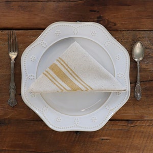 Puede incluir: Un plato de cerámica blanco con borde festoneado y una servilleta de lino a rayas blancas y doradas doblada en forma de triángulo. El plato está sobre una mesa de madera con un tenedor y una cuchara de plata.