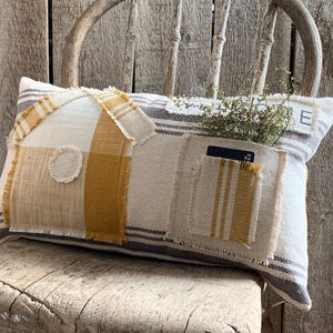 May include: A decorative rectangular pillow with a house-shaped design. The pillow features a patchwork of beige, yellow, and striped fabrics. A small pocket and a circular detail are visible on the front. The pillow is resting on a rustic wooden chair.