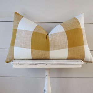 May include: A rectangular pillow with a large buffalo check pattern in mustard yellow and white. The pillow is resting on a small, white, distressed table against a white horizontal plank background. The pillow is a decorative home accent.
