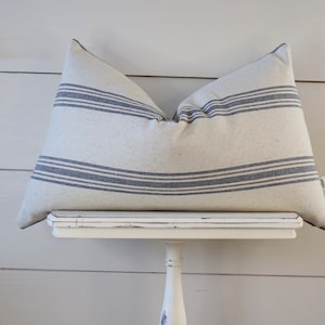 May include: A white and blue striped pillow with a subtle texture. The pillow is sitting on a white pedestal.