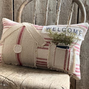 May include: A white and red striped pillow with a pocket and the word "WELCOME" in black lettering. The pillow is sitting on a wooden chair.