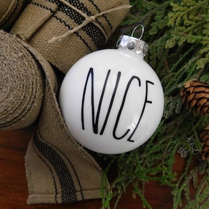 May include: White Christmas ornament with black lettering that reads "NICE". The ornament is surrounded by burlap ribbon and evergreen branches.