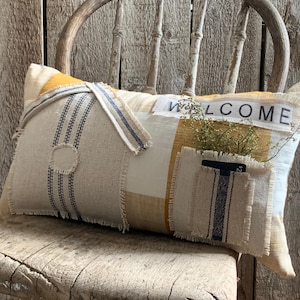 Puede incluir: Una almohada decorativa con un diseño de patchwork que presenta una tela de cuadros beige, blanca y amarilla, una tela beige con rayas negras y blancas y una tela beige con un bolsillo. La almohada tiene la palabra "WELCOME" cosida en hilo negro. La almohada está sentada en una silla de madera.