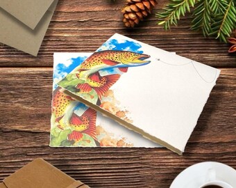 May include: Two blank greeting cards with a watercolor illustration of a fish jumping out of the water. The fish is orange, yellow, and red with blue accents. The cards are on a wooden surface with a pine cone and greenery.