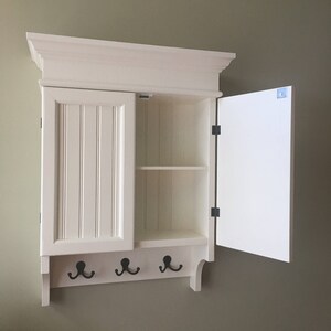 May include: White wall-mounted cabinet with a single door and a shelf inside. The cabinet has a decorative trim and three hooks on the bottom.