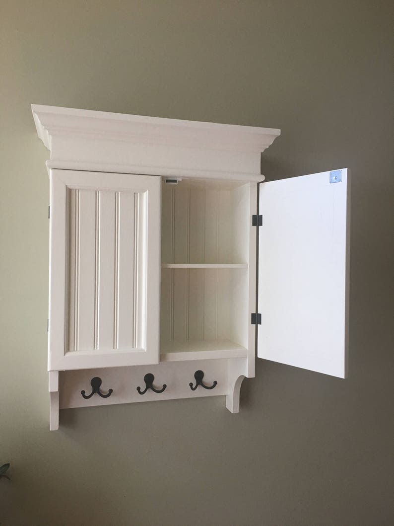 May include: White wall-mounted cabinet with a single door and a shelf inside. The cabinet has a decorative trim and three hooks on the bottom.