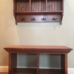 May include: A wooden entryway bench with a shelf and three hooks above. The bench has a dark brown stain and is made of solid wood. The shelf has three compartments and is also stained dark brown.