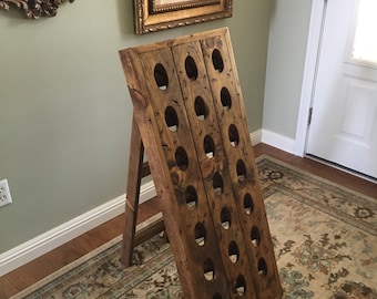 May include: A wooden wine rack with a distressed brown finish. The rack is made of two pieces of wood that are joined together to form a triangular shape. The rack has 20 holes for holding wine bottles. The rack is standing on a patterned rug.