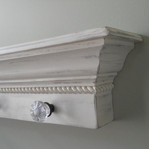 Puede incluir: Estante de madera pintada de blanco con un ribete decorativo de cuerda y tres pomos de cristal. El estante está montado en una pared.