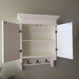May include: White wooden wall mounted cabinet with two doors and three hooks. The doors are open and the cabinet has one shelf.