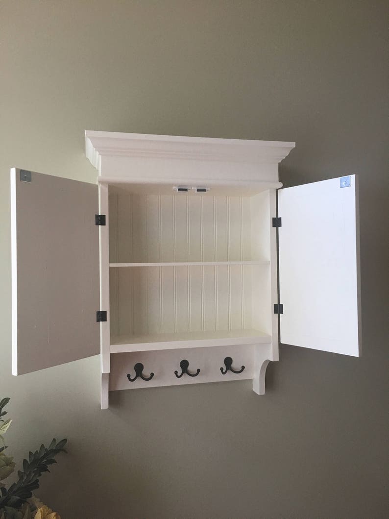May include: White wooden wall mounted cabinet with two doors and three hooks. The doors are open and the cabinet has one shelf.