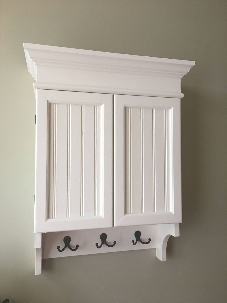 May include: White wall-mounted cabinet with two doors and a shelf. The cabinet has a decorative trim around the top and three hooks on the bottom.
