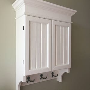 May include: White wall mounted cabinet with two doors and a decorative trim. The cabinet has a shelf and three hooks on the bottom.