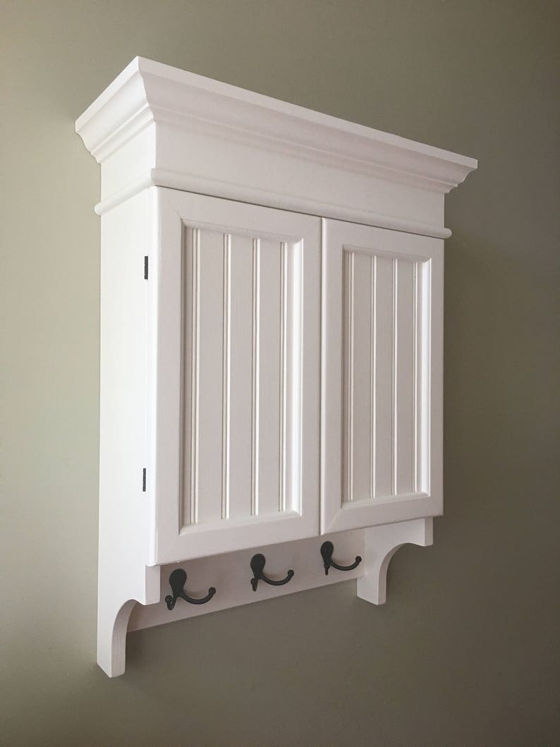 May include: White wall mounted cabinet with two doors and a decorative trim. The cabinet has a shelf and three hooks on the bottom.