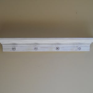 May include: White wooden shelf with decorative trim and four crystal knobs. The shelf is mounted on a wall.