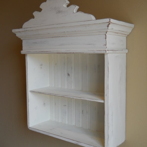 Puede incluir: Estantería de pared de madera pintada de blanco con una parte superior decorativa y dos estantes. La estantería tiene un acabado desgastado.