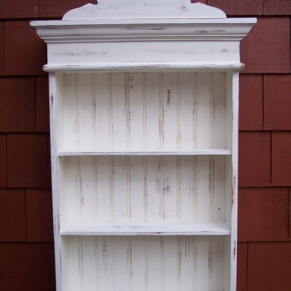 Puede incluir: Estantería de madera blanca con tres estantes y una parte superior decorativa. La estantería tiene un acabado de pintura blanca desgastada.
