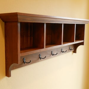 May include: A wooden wall shelf with four cubbies and six hooks. The shelf is stained a dark brown color and has a decorative trim around the top and bottom.