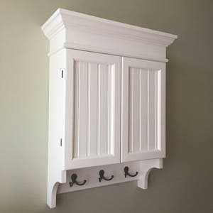 May include: White wall mounted cabinet with two doors and a shelf. The cabinet has a decorative trim around the top and a shelf with four hooks underneath.