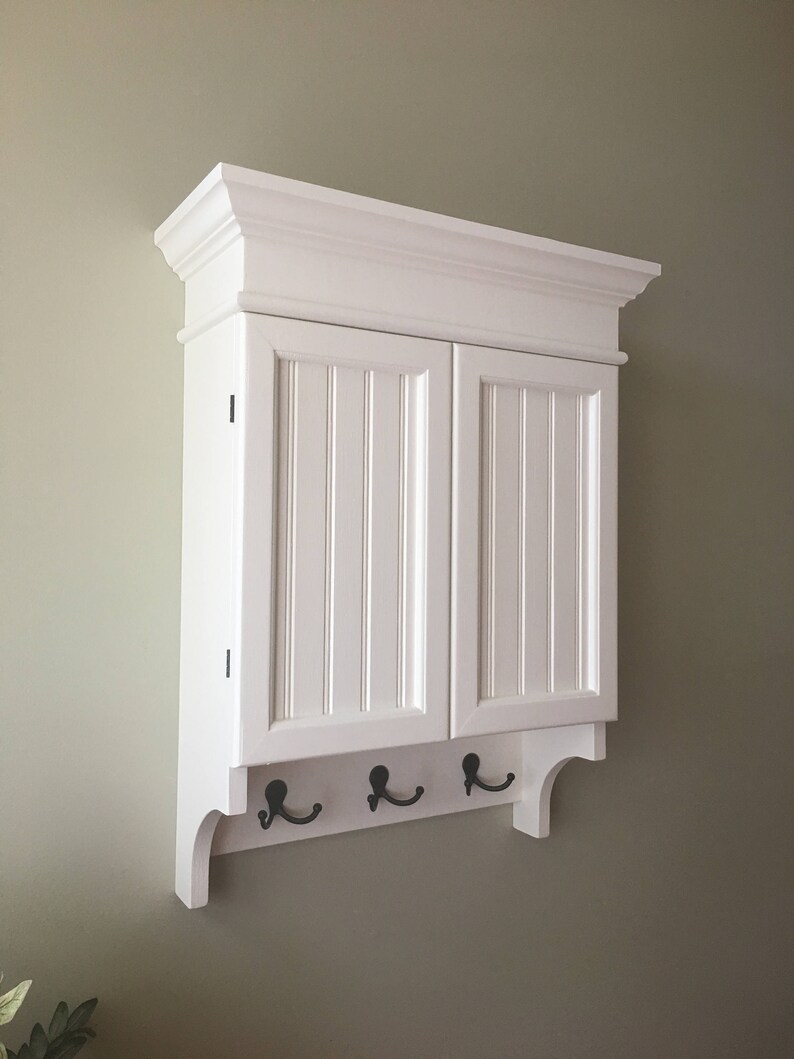 May include: White wall mounted cabinet with two doors and a shelf. The cabinet has a decorative trim around the top and a shelf with four hooks underneath.