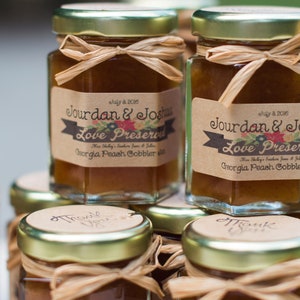 May include: Small glass jars filled with a golden brown substance, tied with twine and decorated with personalized labels that read "Jourdan & Josh Love Preserved Georgia Peach Cobbler".