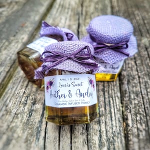 May include: Three small glass jars with purple burlap lids and labels. The label on the front jar reads "April 18, 2021 Love is Sweet Heather & Andrew Miss Shelly's Southern Jams & Jellies Cinnamon Infused Honey".