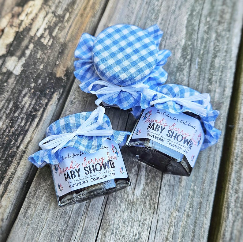 May include: Two small glass jars with blue and white gingham fabric lids. The jars contain blueberry cobbler jam and have labels that say "Thank you for celebrating Mariah's Berry Sweet Baby Shower".