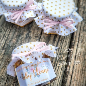 1.5oz Hexagon honey favor with a white personalized label. Fabric topper is White with Gold Polka Dots and a Light Pink raffia ribbon. The label design is our Watercolor Floral has a gold font.