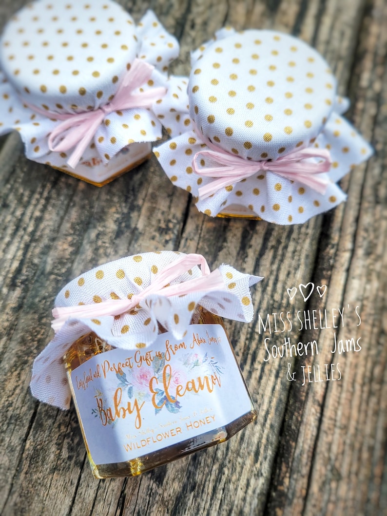 1.5oz Hexagon honey favor with a white personalized label. Fabric topper is White with Gold Polka Dots and a Light Pink raffia ribbon. The label design is our Watercolor Floral has a gold font.