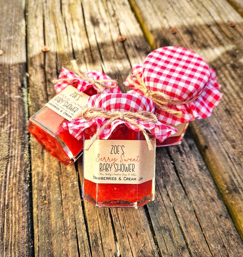 May include: Three small glass jars filled with red jam, each with a red and white checkered fabric lid and a brown paper label. The label on the front jar reads "ZOE'S Berry Sweet BABY SHOWER Strawberries & Cream Jam".