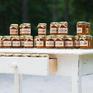 May include: A white distressed wooden table with a drawer, holding small glass jars filled with a golden liquid. The jars are arranged in rows and are topped with lids.