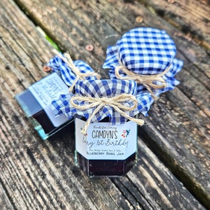May include: Two small glass jars of blueberry basil jam, decorated with blue and white gingham fabric and twine bows. The labels read "Thanks for Coming, Camdyn's 1st Birthday, Blueberry Basil Jam".