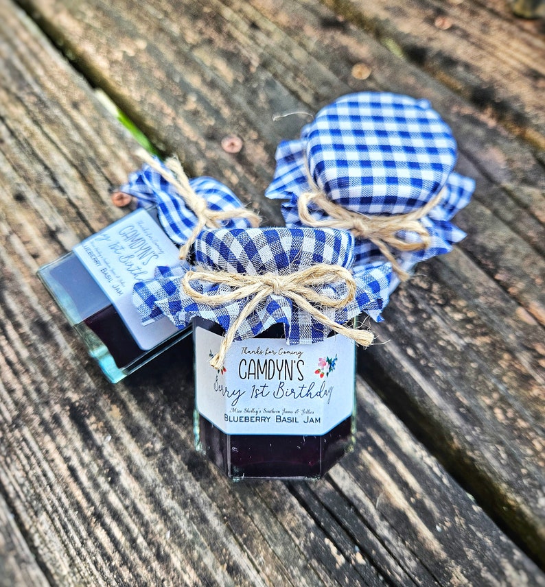May include: Two small glass jars of blueberry basil jam, decorated with blue and white gingham fabric and twine bows. The labels read "Thanks for Coming, Camdyn's 1st Birthday, Blueberry Basil Jam".